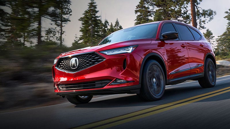 Red Acura MDX SUV driving on a paved road with blurred evergreen trees in the background.