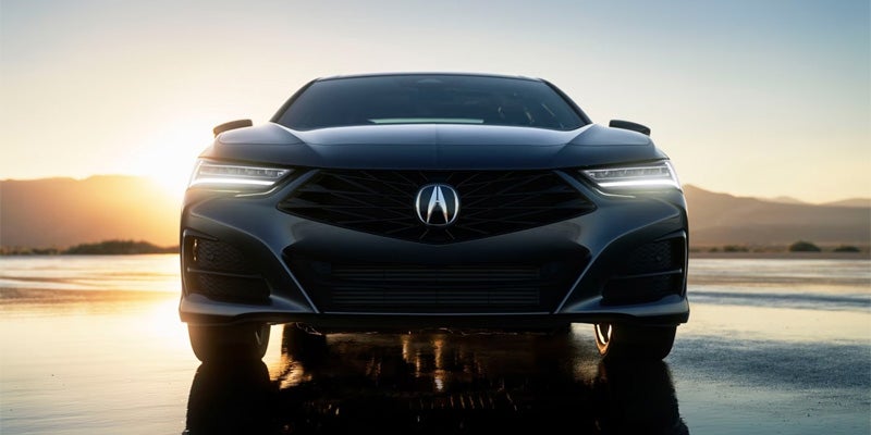 Front view of a dark Acura sedan on a wet reflective surface at sunset, with illuminated headlights and mountains in the distance