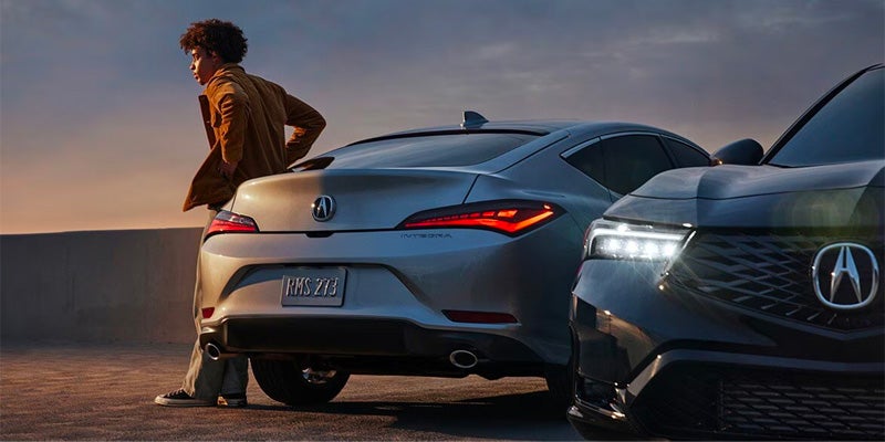 White Acura hatchback parked beside a dark-colored Acura SUV at sunset, viewed from the rear