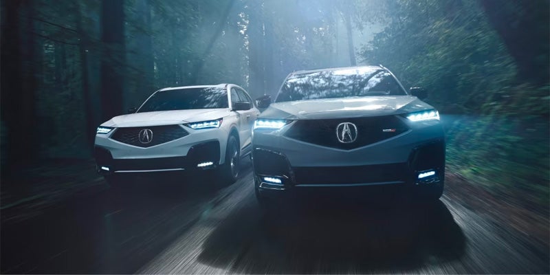 Two Acura SUVs driving side by side through a forested road, featuring bright LED headlights