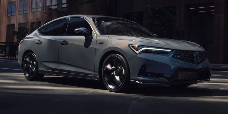 Silver Acura sedan driving on a city street, shown from a front three-quarter angle with black alloy wheels and modern LED headlights