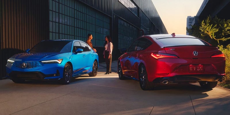 Blue Acura sedan and red Acura sedan parked outside a modern building, viewed during daylight with two people standing between the vehicles