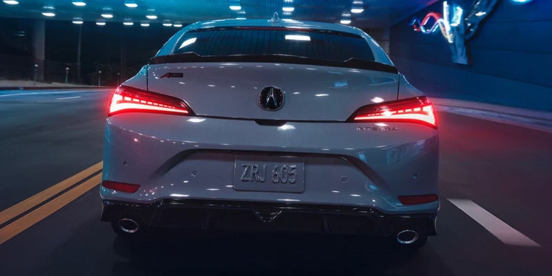 Rear view of a white Acura sedan driving through a brightly lit tunnel at night, with illuminated LED taillights and dual exhaust outlets visible
