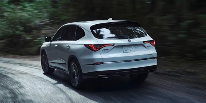 White Acura SUV driving on a winding road through a forested area, shown from a rear three-quarter angle