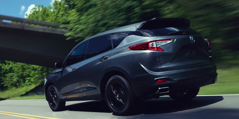 Rear three-quarter view of a dark gray Acura SUV driving on a road beneath an overpass