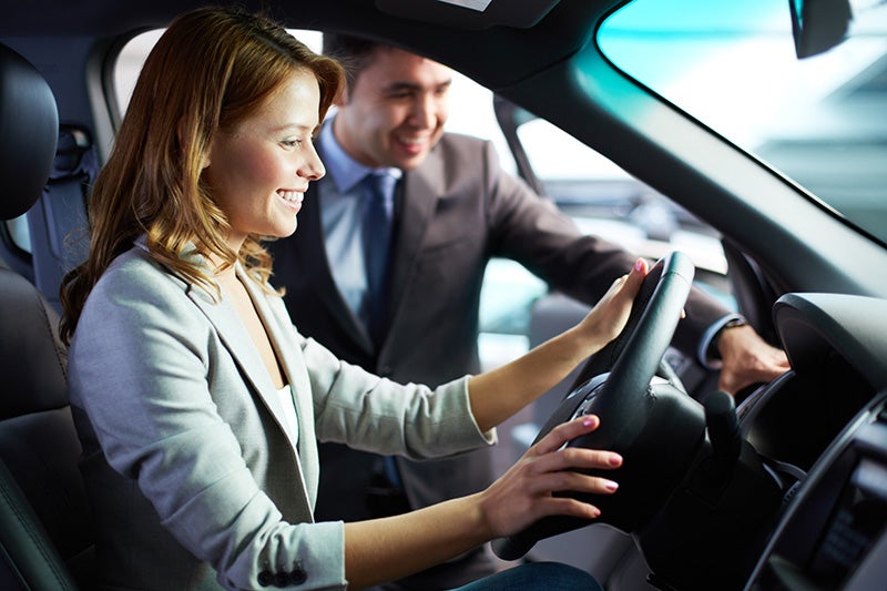 Smiling woman in driver's seat holding the steering wheel, talking to a smiling car salesperson leaning into the car.