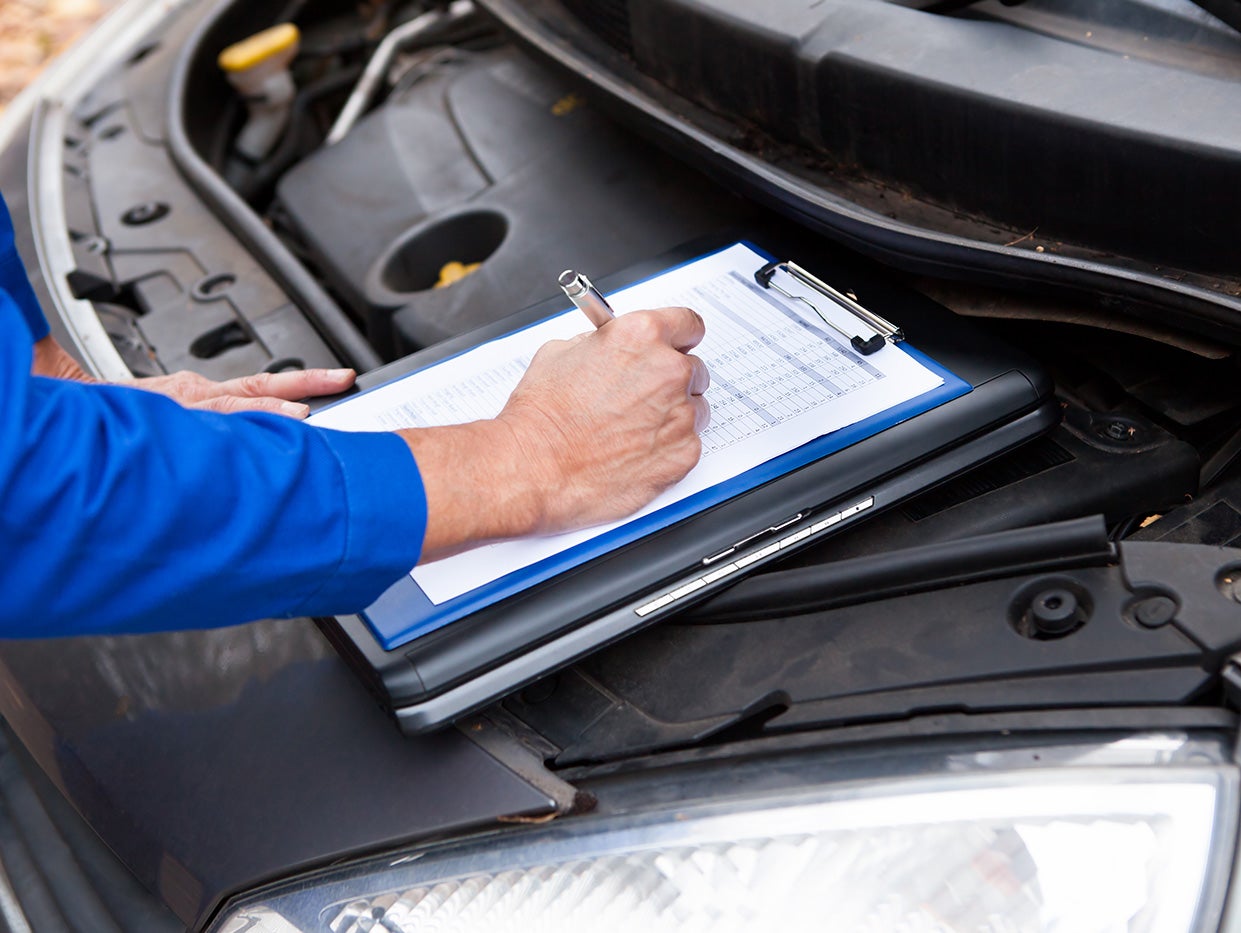 Mechanic in a blue uniform writing on a checklist clipboard over an open car engine.